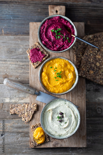 Three hummus dips, beet hummus, sweet potato or carrot or pumpkin hummus and parsnip hummus with flax seeds crackers on a rustic wooden board with a purple napkin on a light natural stone. Top view