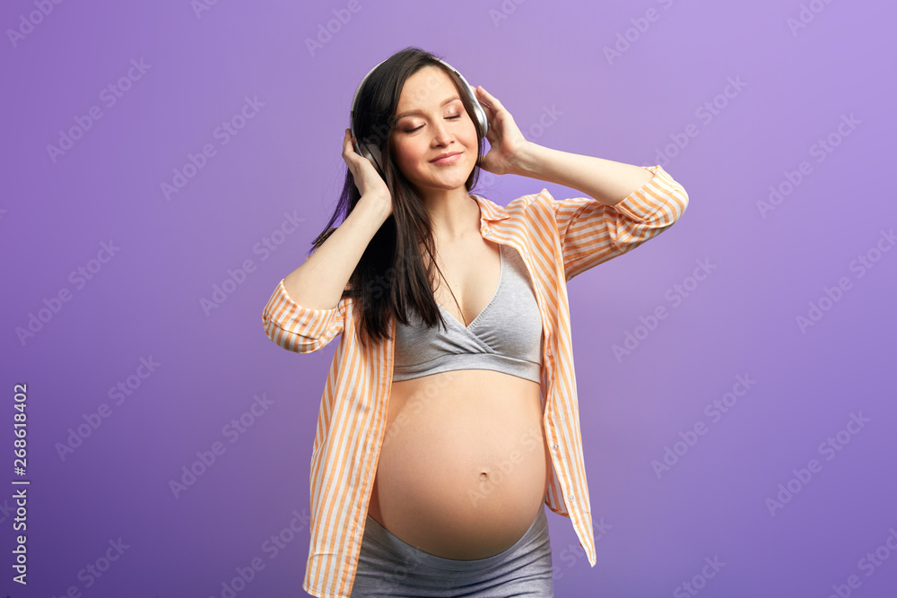 Fototapeta premium Happy pregnant woman feeling healthy listening to music in headphones, dancing in studio over purple background