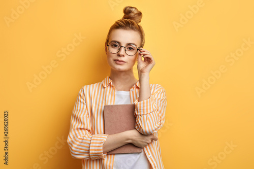 clever cute awesome woman holding a book, touching her glasses and posing to the camera. isolated yellow background, studio shot.