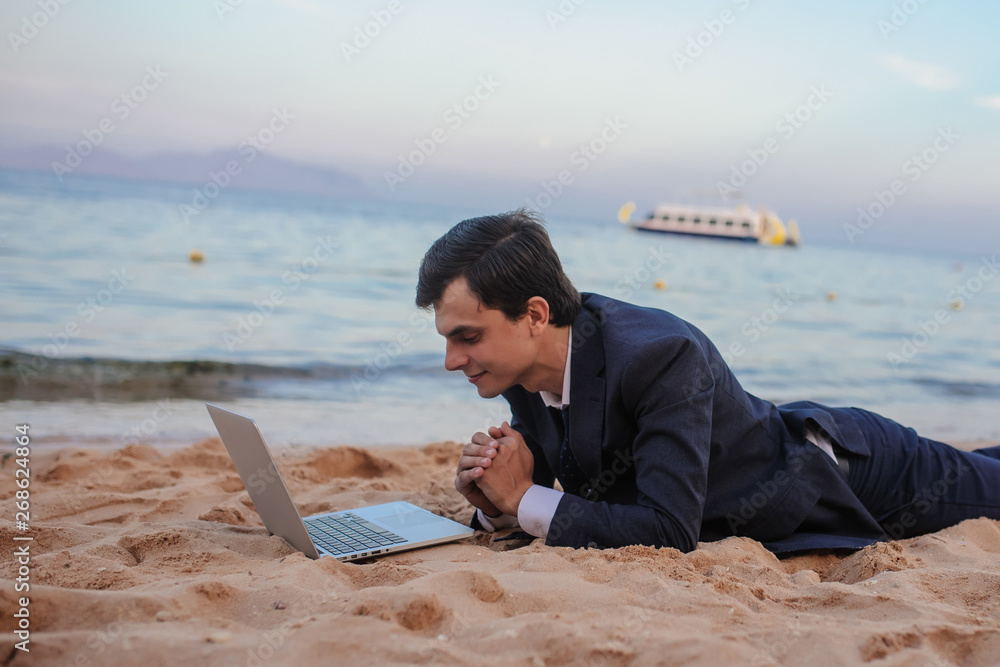 close up photo of a young man in suit with laptop lies on the beach and talking to someone on computer