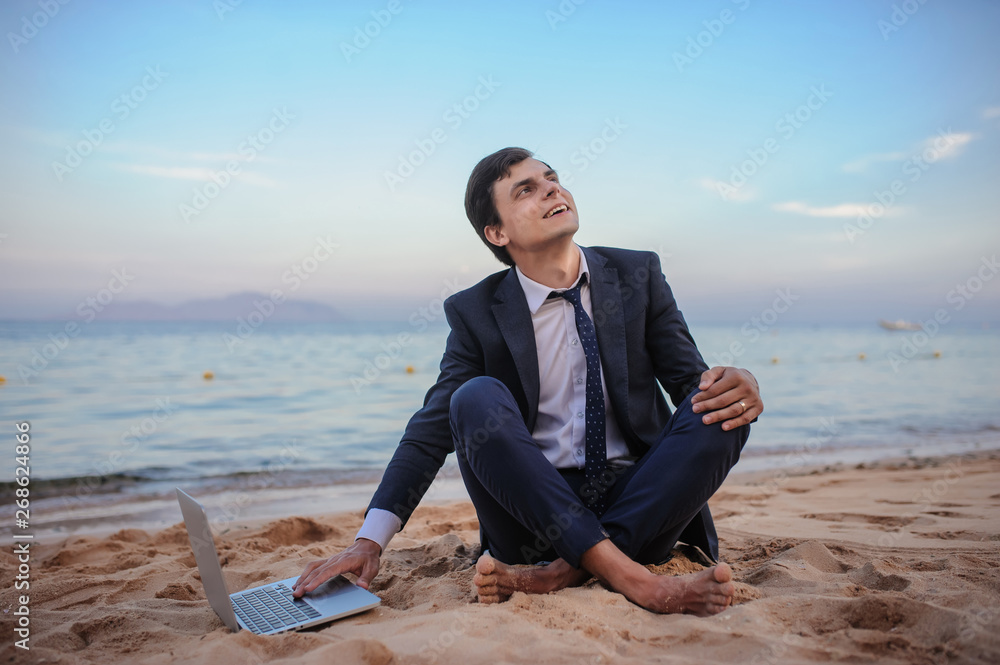lateral view of a young man in suit with laptop sitting on the beach working on the seaside lateral view of a young man in suit with laptop sitting on the beach working on the seaside
