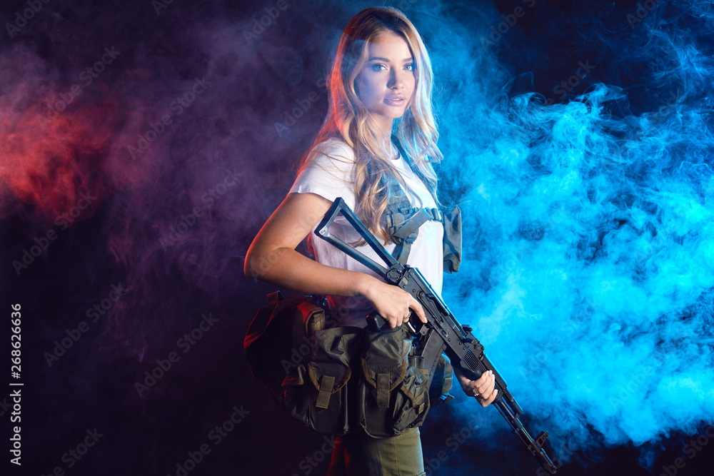 Young blonde female snipper in military outfit with assault rifle in studio on smoky dark background. Women in military service concept.