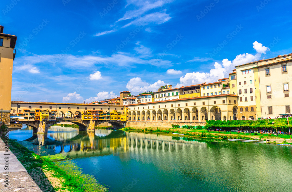 Ponte Vecchio bridge with colourful buildings houses over Arno River blue reflecting water and embankment promenade archways, historical centre of Florence city, blue sky white clouds, Tuscany, Italy