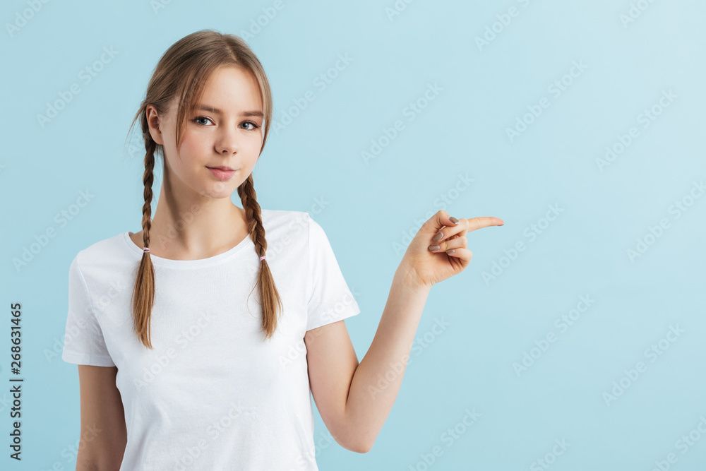 Young beautiful girl with two braids in white t-shirt dreamily looking in camera showing with index finger at empty space over blue background isolated Young beautiful girl with two braids in white t-shirt dreamily looking in camera showing with index finger at empty space over blue background isolated