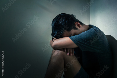 Young depressed asian man sitting alone in dark room with low light environment, PTSD Mental health concept, Selective focus.