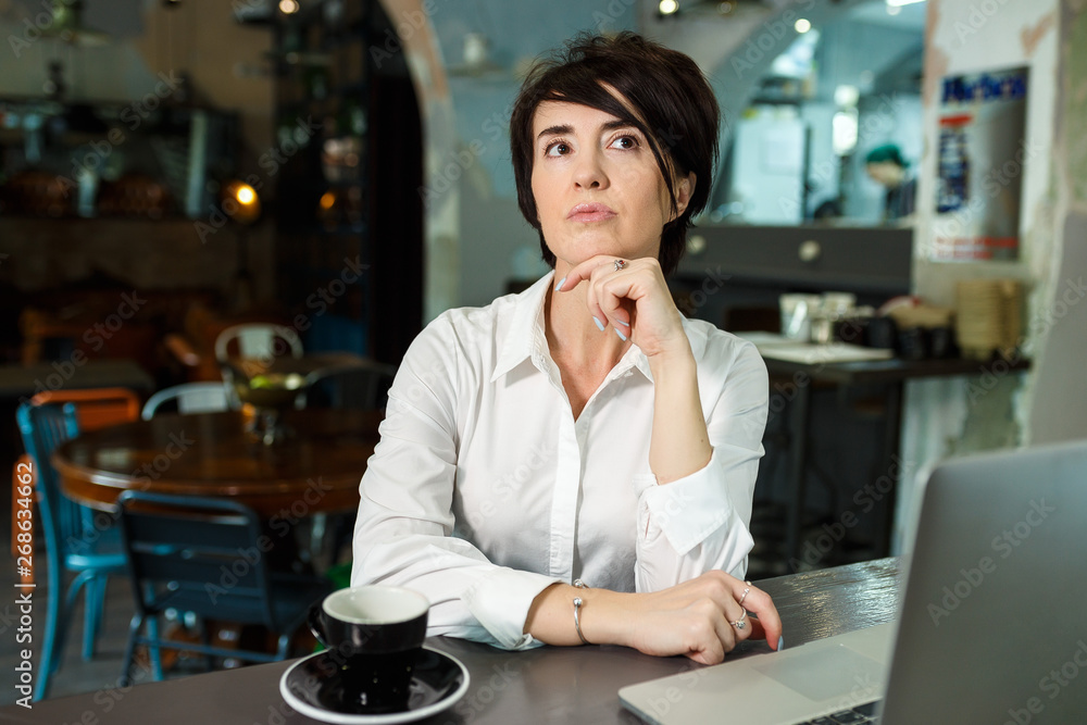 A middle-aged woman sits in a cafe, drinks coffee and works at a computer. A woman is thinking about something A middle-aged woman sits in a cafe, drinks coffee and works at a computer. A woman is thinking about something