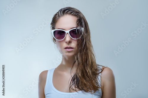 Close up portrait of beautiful young woman in fashionable white sunglasses and white shirt is posing stylishly isolated on gray background in a studio