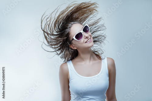 Portrait of happy young woman in fashionable white sunglasses and white shirt with flying hair isolated on gray background in a studio. Fashion accessories photography with beautiful girl