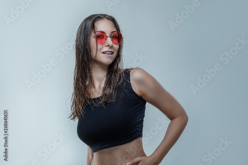Beautiful young woman in fashionable round pink sunglasses and black shirt with wet hair isolated on gray background in a studio. Sexy slim girl is smiling and posing stylishly 
