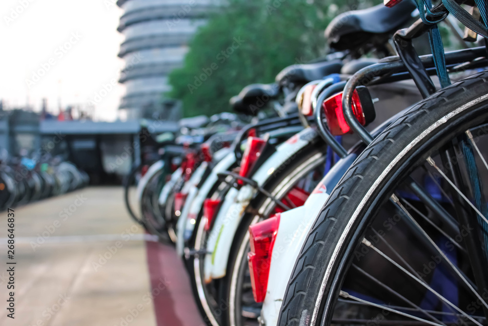 Urban shot of a close-up bicycle wheel with bicycle parking on the background of high-rise building facades and green trees in the back. Urban shot of a close-up bicycle wheel with bicycle parking on the background of high-rise building facades and green trees in the back.