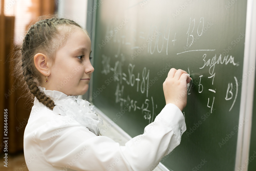 A girl female elementary school student writes on the blackboard. little schoolgirl holds a chalk in her hand and writes a solution to the problem on the school blackboard. A girl female elementary school student writes on the blackboard. little schoolgirl holds a chalk in her hand and writes a solution to the problem on the school blackboard.