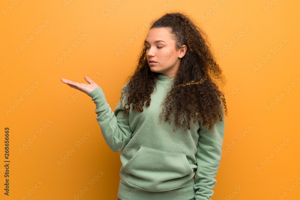 Teenager girl over ocher wall holding copyspace with doubts Teenager girl over ocher wall holding copyspace with doubts