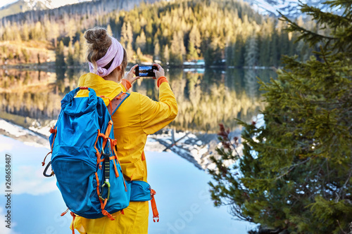Back view of active female tourist photographs lakescape with mountains on her smart phone device, wears yellow raincoat, has blue backpack, takes picture of majestic nature during sunny day.