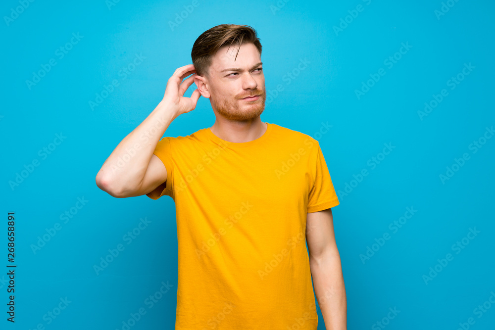 Redhead man over blue wall having doubts while scratching head Redhead man over blue wall having doubts while scratching head