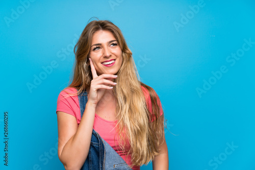Young blonde woman with overalls over isolated blue wall thinking an idea while looking up