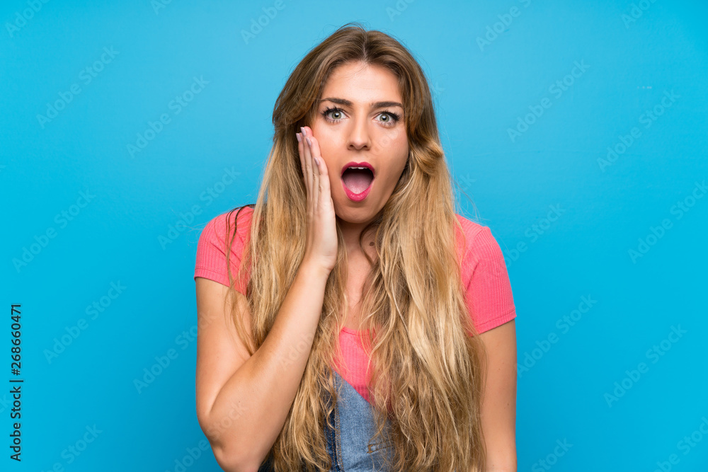 Young blonde woman with overalls over isolated blue wall with surprise and shocked facial expression