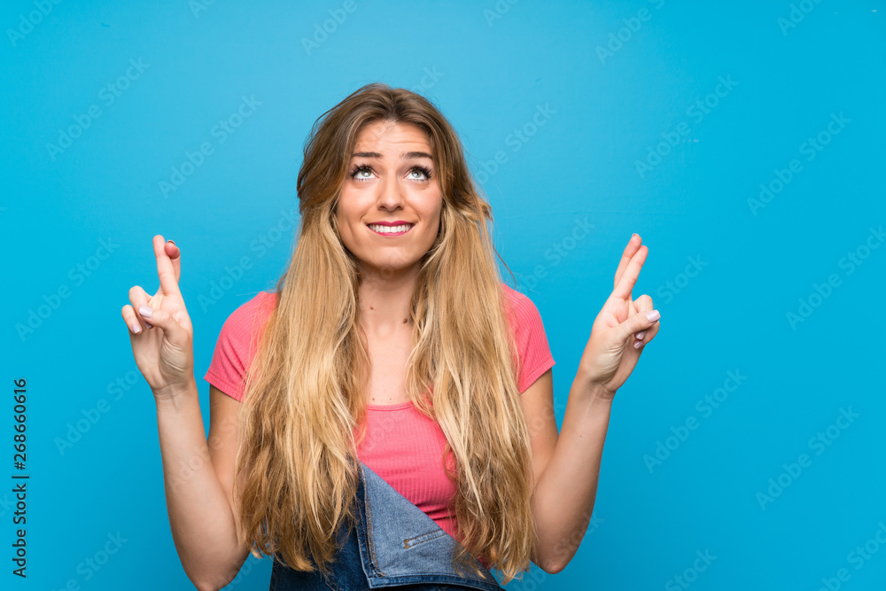 Young blonde woman with overalls over isolated blue wall with fingers crossing and wishing the best Young blonde woman with overalls over isolated blue wall with fingers crossing and wishing the best