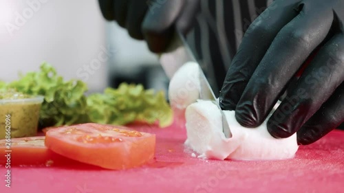 Closeup hands of male professional chef in black gloves are slicing mozzarella cheese on plastic red board in restaurant. photo