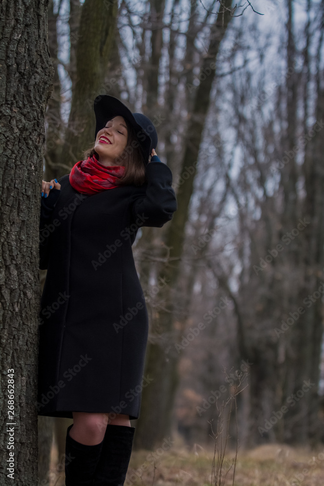 The girl in a black coat, a shovel hat and with red lipstick on lips, walks in the park. Style and modern fashion. The girl in a black coat, a shovel hat and with red lipstick on lips, walks in the park. Style and modern fashion.