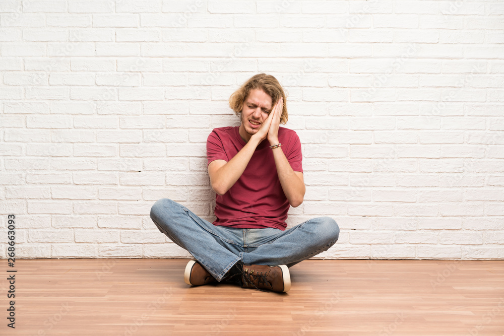 Blonde man sitting on the floor making sleep gesture in dorable expression Blonde man sitting on the floor making sleep gesture in dorable expression
