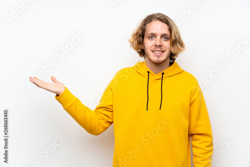 Blonde man with sweatshirt over white wall holding copyspace imaginary on the palm to insert an ad
