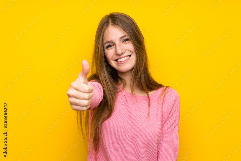 Young woman with long hair over isolated yellow wall with thumbs up because something good has happened Young woman with long hair over isolated yellow wall with thumbs up because something good has happened