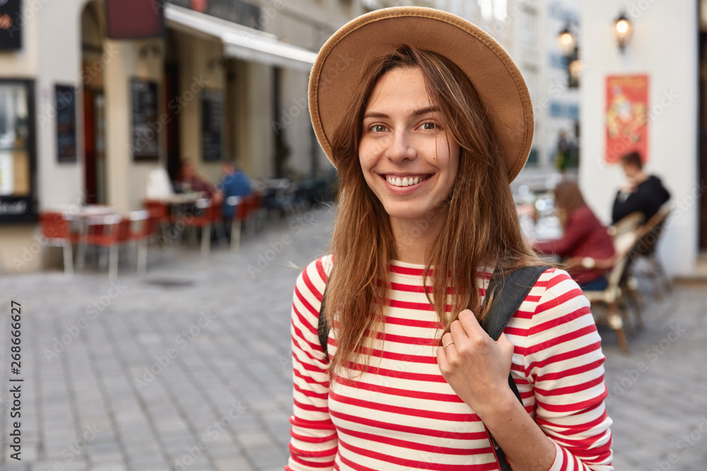 Φωτογραφία Shot of lovely female smiles gladfully, wears hat and striped jumper, being in good mood as strolls across city, has excursion during trip, poses outdoor, has spare time