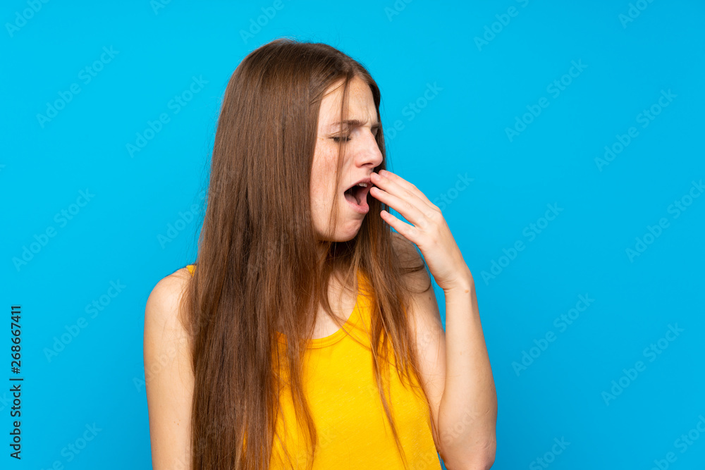 Young woman with long hair over isolated blue wall yawning and covering wide open mouth with hand Young woman with long hair over isolated blue wall yawning and covering wide open mouth with hand