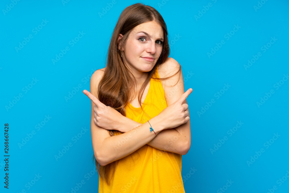 Young woman with long hair over isolated blue wall pointing to the laterals having doubts Young woman with long hair over isolated blue wall pointing to the laterals having doubts