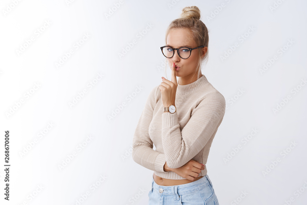 Shhh sweety. Indoor shot of feminine sensual and sexy blond woman in glasses and combed hair showing shush sign with index finger over folded lips hiding mystery over white background Shhh sweety. Indoor shot of feminine sensual and sexy blond woman in glasses and combed hair showing shush sign with index finger over folded lips hiding mystery over white background