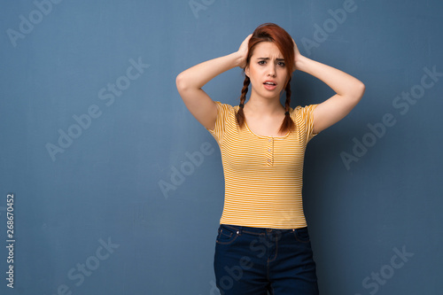 Young redhead woman over blue background takes hands on head because has migraine