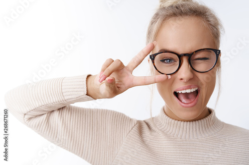 Hey smile you. Portrait of cheeky and cute glamour blond woman in glasses combed hair and sweater winking happily showing peace victory gesture as fooling around, being optimistic