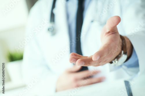Male doctor making welcome gesture  politely inviting patient to sit down in medical office. Photo with depth of field.