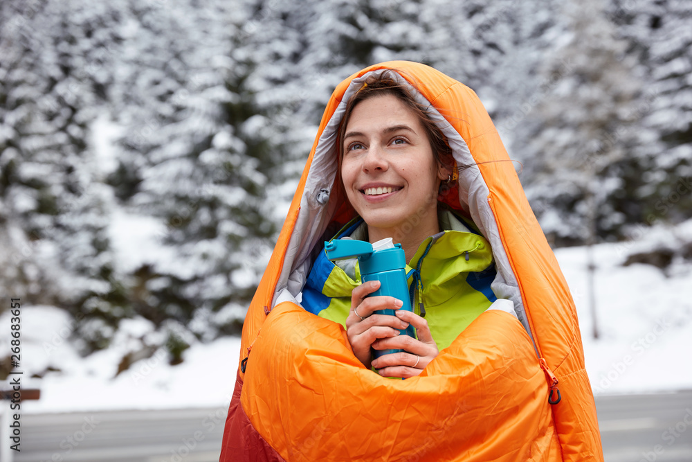 Φωτογραφία Dreamy positive woman reaches top of mountains, wrapped in sleeping bag during cold weather, drinks hot drink from flask, poses over beautiful snowy view, focused into distance, being happy