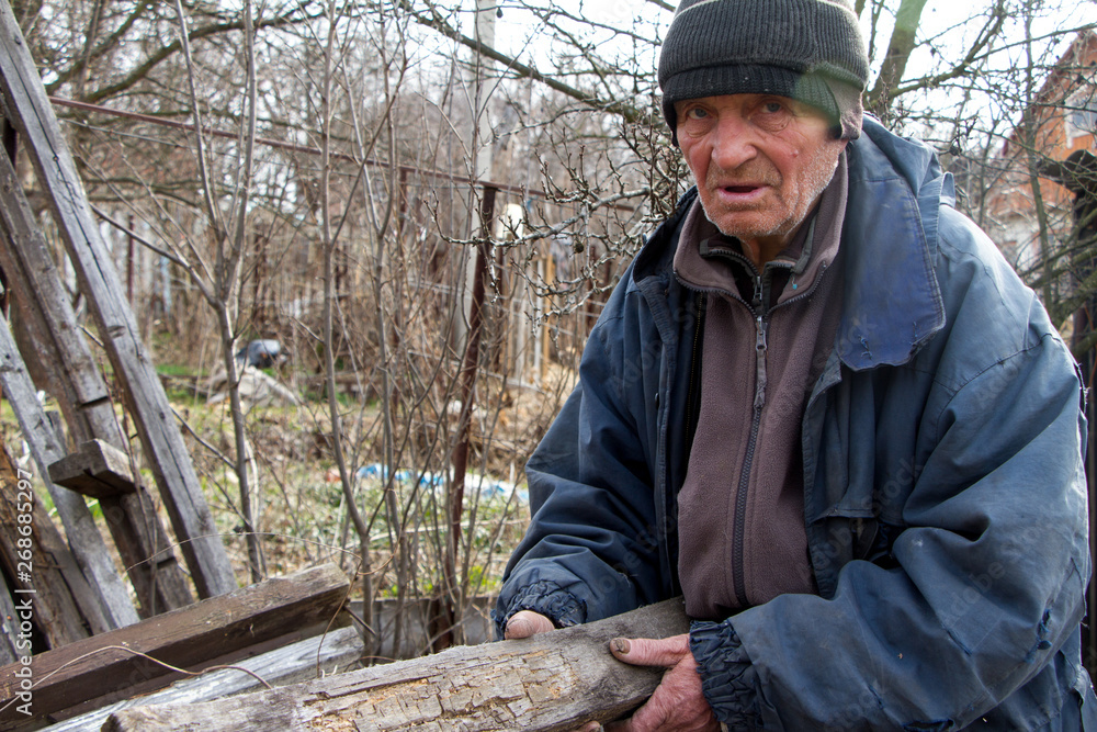Obraz premium An old man in untidy clothes chooses from a pile of old wood boards to heat the house, loneliness in the village