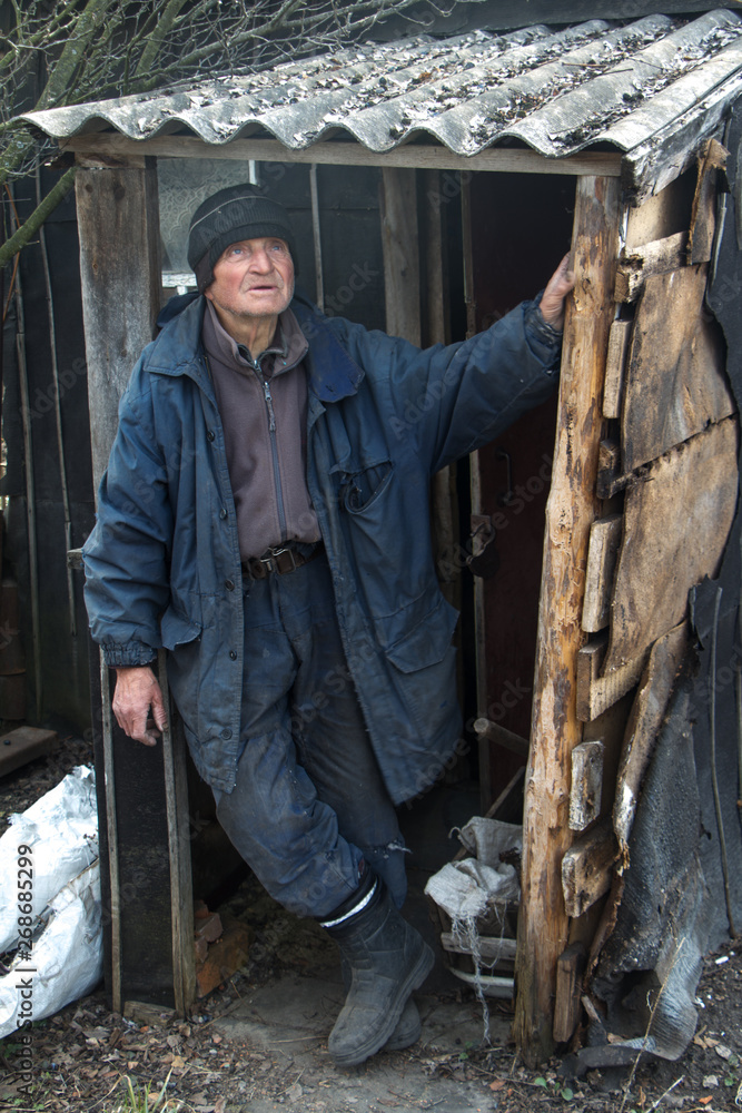 An old man in sloppy clothes stands on the threshold of his own ruined house and looks into the distance An old man in sloppy clothes stands on the threshold of his own ruined house and looks into the distance