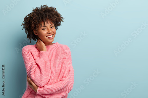 Studio shot of glad charming young female with Afro haircut, touches neck, wears oversized jumper, isolated over blue background with blank space for your promotional content. Pleasant emotions photo