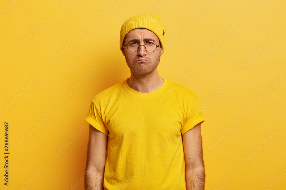 Unhappy unsatisfied guy smirks face, has gloomy expression, wears yellow hat and t shirt, puzzled by bad news, poses over studio wall, being offended by someone. Negative human emotions and feelings