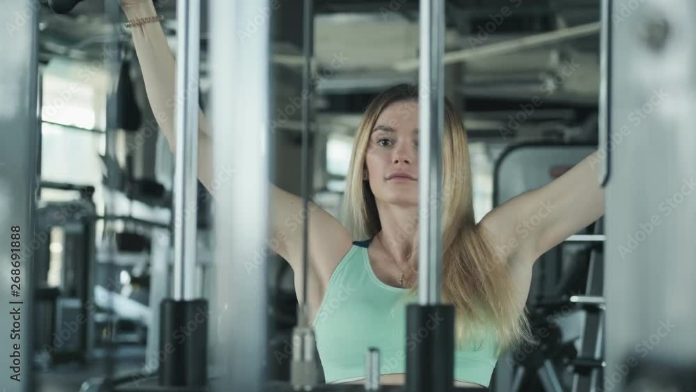Fitness blonde girl doing exercise for shoulders. Young woman lifting weights in gym. Medium shot of blonde female in fitness center in slow motion. Fitness blonde girl doing exercise for shoulders. Young woman lifting weights in gym. Medium shot of blonde female in fitness center in slow motion.