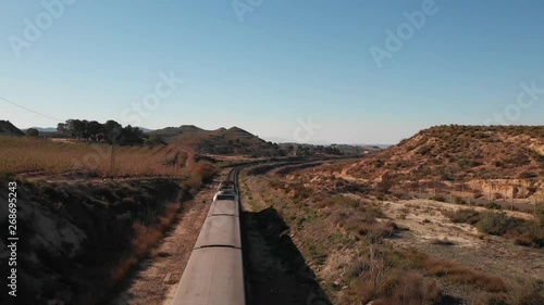 The country of Murcia in Spain in the late afternoon in spring. A passenger train drives through a long bend and then under a road bridge south to Murcia. The sun is shining. Aerial view HD.