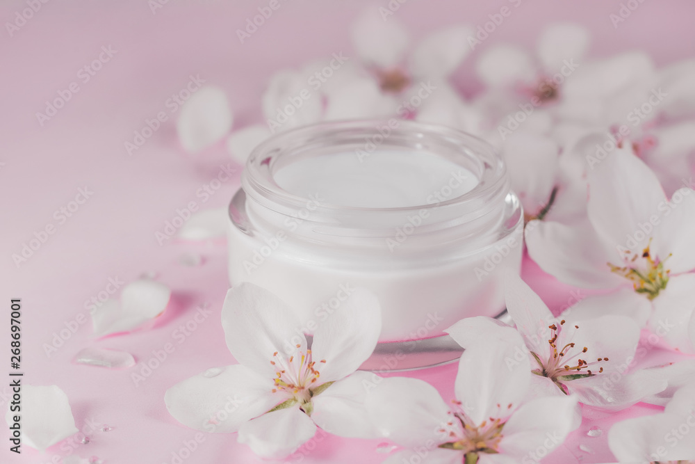 Beauty concept. Jar of face cream surrounded by blossoming spring flowers on pink background close up.