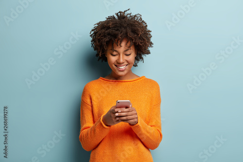 Satisfied hipster girl with Afro haircut, types text message on cell phone, enjoys online communication, types feedback, wears orange jumper, isolated on blue studio wall. Technology concept photo