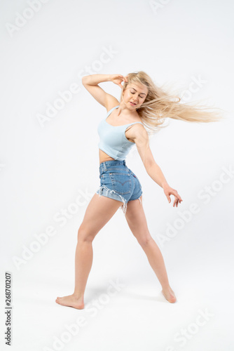 Beautiful girl in a blue T-shirt and denim shorts is dancing on a white background. Isolated. The girl has long blond hair