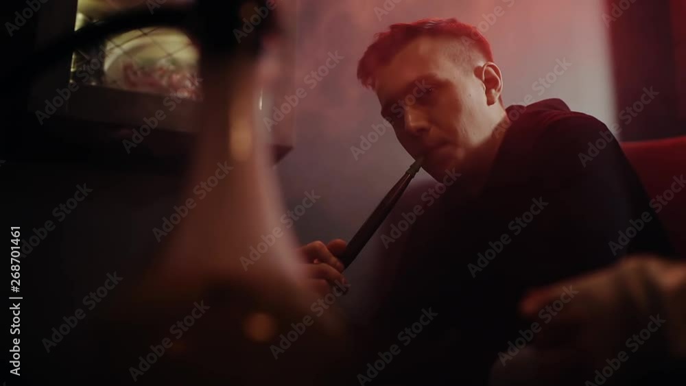 Close-up beautiful guy with the fashionable hairstyle smoking hookah and exhales smoke through his nose and mouth at dark room of shisha lounge in slow motion. Young hipster man relaxing with friends Close-up beautiful guy with the fashionable hairstyle smoking hookah and exhales smoke through his nose and mouth at dark room of shisha lounge in slow motion. Young hipster man relaxing with friends