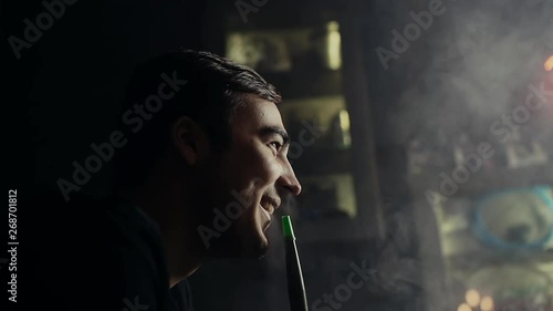 Close-up handsome guy smokes hookah and has fun with his friendsat dark room of shisha lounge in slow motion. Young hipster man smoking a hookah and exhales with a smile through his mouth photo