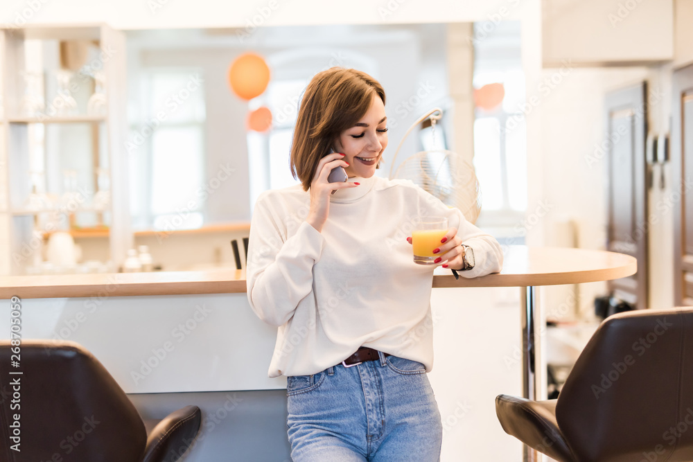 Smiling young woman using mobile phone, holding glass of orange juice at the kitchen Smiling young woman using mobile phone, holding glass of orange juice at the kitchen