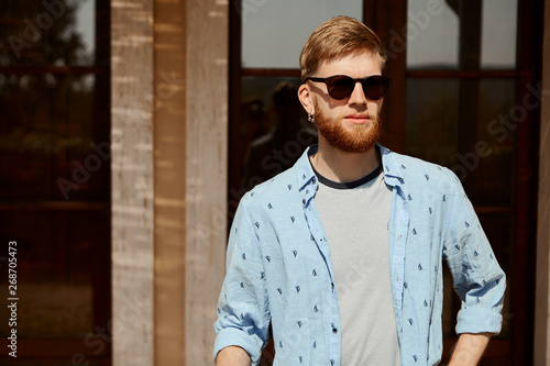 People, summertime, style and fashion concept. Attractive young guy with thick beard and pierced ears posing against blurred house background looking at camera wearing stylish black sunglasses