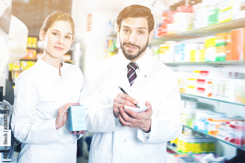 Two pharmacists are inventorying medicines with note near shelves in apothecary.