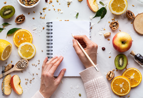 Female hand write in notebook on healthy food background, women diet nutrition recipe menu, fresh summer fruit granola seeds on white table organic super food, health care detox, top view, copy space photo