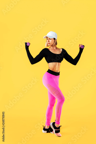 Sporty woman in fashionable sportswear does the exercises with dumbbells. Photo of muscular woman on yellow background. Strength and motivation.